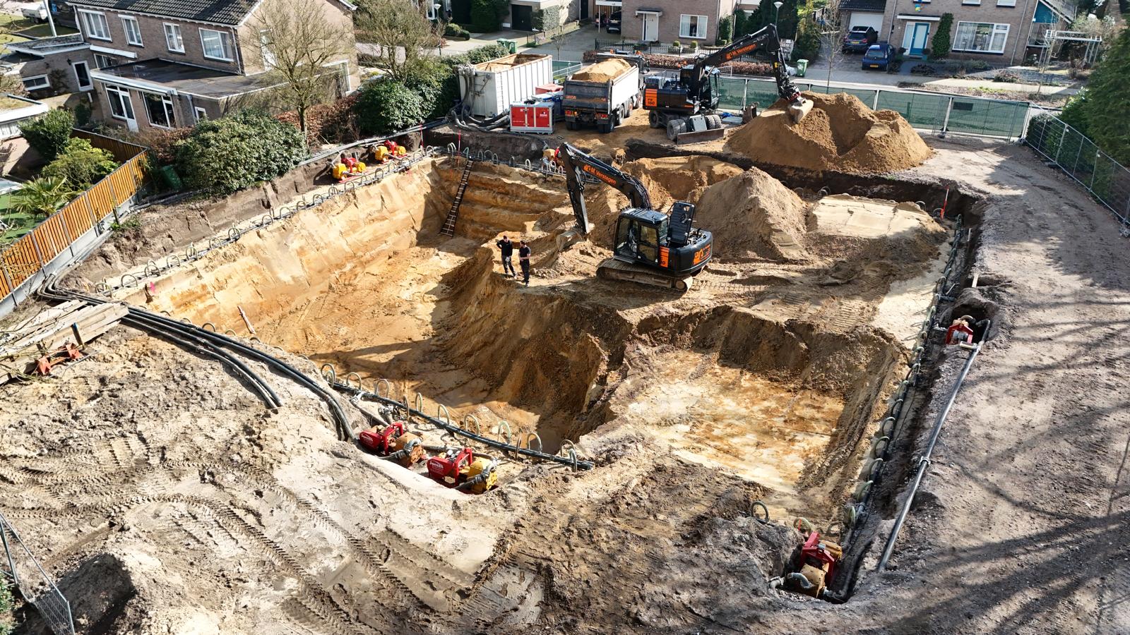 Diepe bouwput in woonwijk met actieve bronbemaling, graafmachines en pompen voor verlaging van het grondwaterpeil tijdens bouwwerkzaamheden.