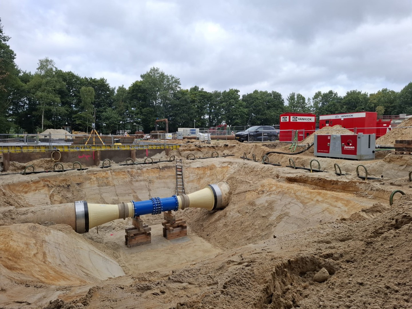 Bouwput met grote ondergrondse leiding op steunblokken, omgeven door zand, machines en rode bouwcontainers op een werkterrein.
