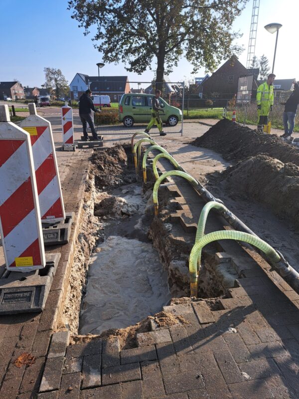 Sleuf in woonstraat met actieve bronbemaling, afzuigslangen en tijdelijke afzetting voor veilig werken aan ondergrondse infrastructuur.